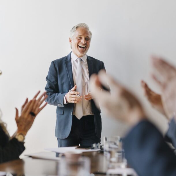 Successful senior businessman smiling and engaging with colleagues during a meeting, with hands applauding, reflecting a positive and collaborative atmosphere in a professional setting.