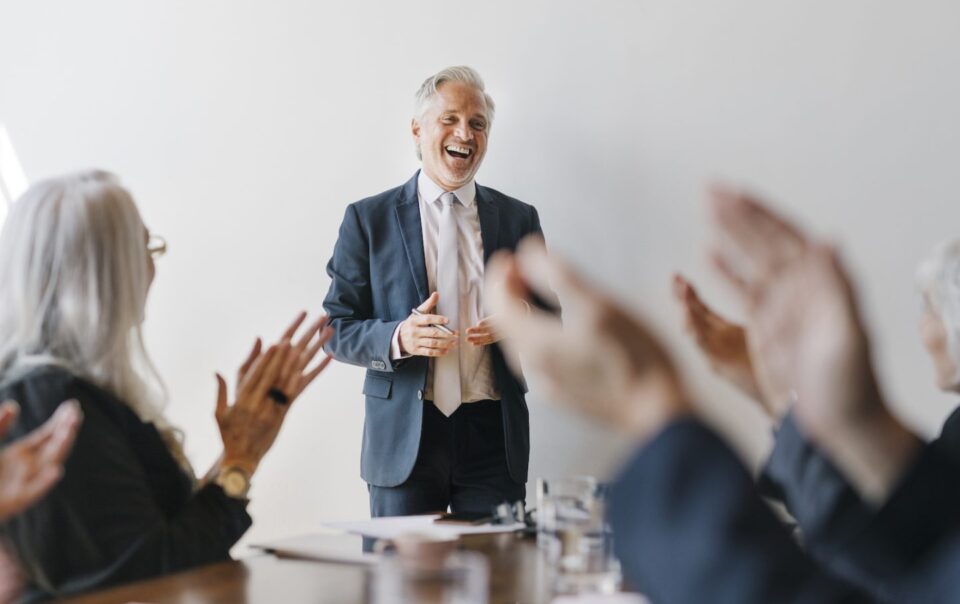 Successful senior businessman smiling during a meeting, receiving applause from colleagues, illustrating positive client interactions in financial legal services.